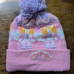 May include: Pink knit beanie with a purple pom-pom. The hat features a repeating pattern in pink, white, and yellow, with a Cabbage Patch Kids logo on the folded brim. The hat is made of a soft, knitted material.