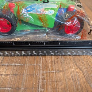 May include: A green toy car with red wheels is packaged in a clear plastic bag. The car has blue and white accents and the text "COLLATI" is visible. A ruler is placed below the car, showing measurements in centimetres.