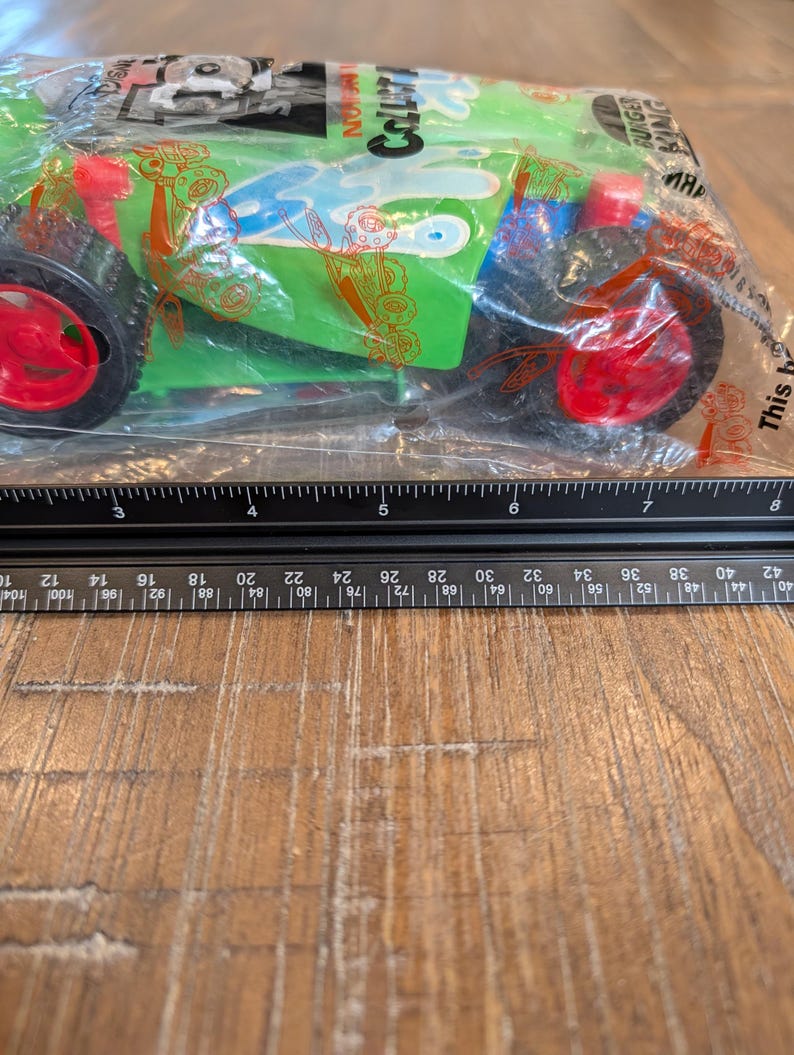 May include: A green toy car with red wheels is packaged in a clear plastic bag. The car has blue and white accents and the text "COLLATI" is visible. A ruler is placed below the car, showing measurements in centimetres.