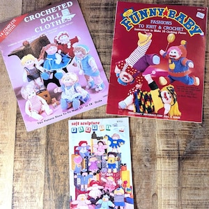 May include: Three vintage craft books on a wooden surface. The books are titled "Crocheted Doll Clothes", "Funny Baby Fashions to Knit & Crochet", and "Soft Sculpture Playtime". The covers feature illustrations of dolls and knitted clothing.