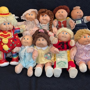 May include: A collection of Cabbage Patch Kids dolls, each with unique features. The dolls have various hair colors, outfits, and accessories, including a clown, a football player, and dolls in dresses. The dolls are arranged on a dark blue surface.