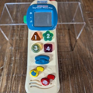 May include: A cream-colored VTech Little Smart Tiny Touch Phone Plus toy with a blue screen and buttons. The phone has colorful buttons with numbers and musical notes. A red knob is at the top.