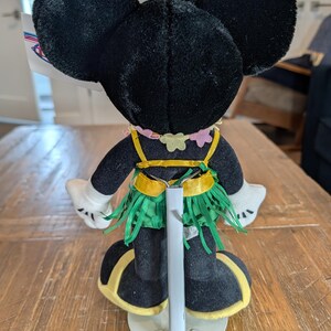 May include: A plush Minnie Mouse doll, viewed from the back, wearing a Hawaiian-themed outfit. The doll has black ears, a pink and yellow lei, a green grass skirt, and yellow accents on her shoes. The doll is displayed on a wooden table.