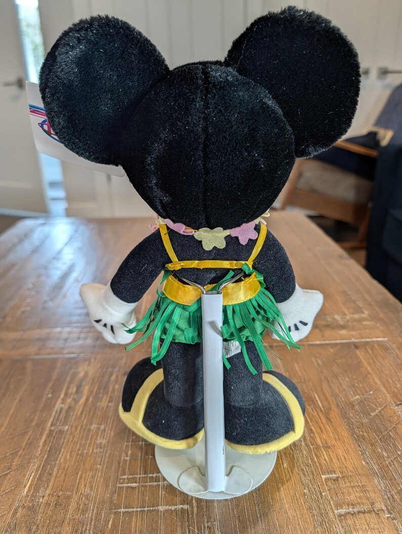 May include: A plush Minnie Mouse doll, viewed from the back, wearing a Hawaiian-themed outfit. The doll has black ears, a pink and yellow lei, a green grass skirt, and yellow accents on her shoes. The doll is displayed on a wooden table.