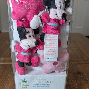 May include: A Disney baby musical mobile in a clear plastic package. The mobile features two Minnie Mouse plush figures in pink dresses and two pink star-shaped plush toys. The packaging includes the text "Sitting Pretty Musical Mobile".