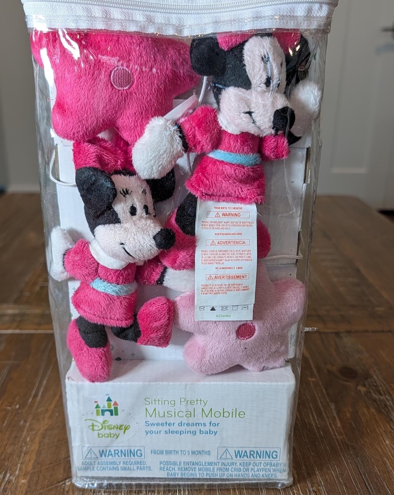 May include: A Disney baby musical mobile in a clear plastic package. The mobile features two Minnie Mouse plush figures in pink dresses and two pink star-shaped plush toys. The packaging includes the text "Sitting Pretty Musical Mobile".