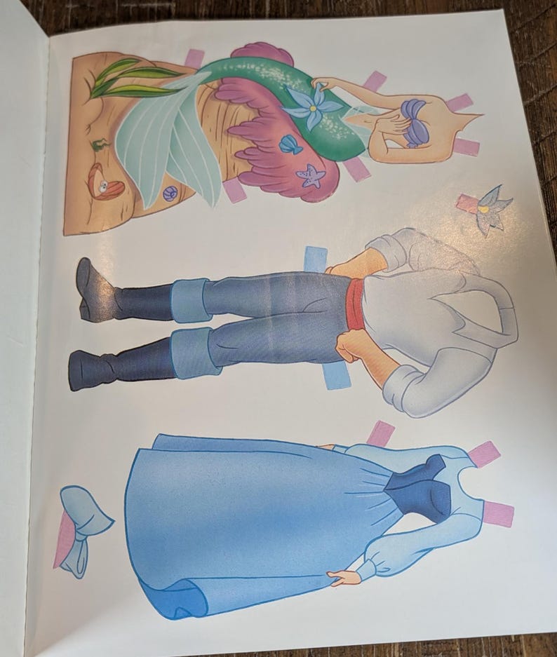 May include: A page from a children's paper doll book featuring three cut-out figures. The figures include a mermaid with a teal tail, a man in blue pants and a white shirt, and a woman in a blue dress. The figures have tabs for easy assembly.