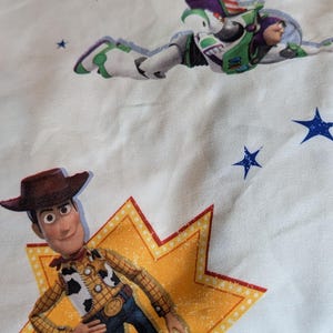 May include: A white fabric featuring a repeating pattern of Toy Story characters including Woody, a cowboy figure standing on a yellow star, and Buzz Lightyear, a space ranger figure in flight, interspersed with small blue stars and planets.