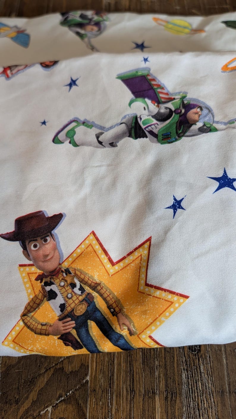 May include: A white fabric featuring a repeating pattern of Toy Story characters including Woody, a cowboy figure standing on a yellow star, and Buzz Lightyear, a space ranger figure in flight, interspersed with small blue stars and planets.