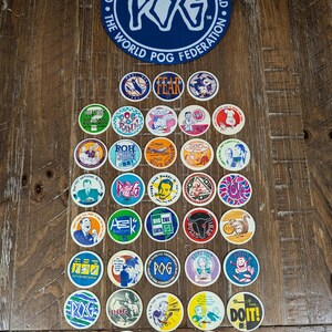 May include: A collection of circular cardboard POG game discs featuring various colorful graphic illustrations and text, arranged below a large blue circular Official Tournament Game Pad from the World POG Federation with white text.