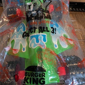 May include: A green and blue toy car from Burger King, in its original packaging. The toy has black wheels with red details. The packaging displays the text "TOY ON VIDEO" and "COLLECT ALL 3!"