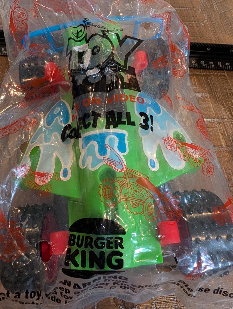 May include: A green and blue toy car from Burger King, in its original packaging. The toy has black wheels with red details. The packaging displays the text "TOY ON VIDEO" and "COLLECT ALL 3!"