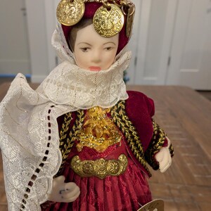 May include: A detailed doll in traditional costume, featuring a burgundy dress and a white lace shawl. The doll has a gold chain headpiece with gold coin accents. The doll's outfit is trimmed with gold and black detailing.