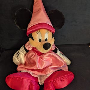 Peut inclure: Poup&eacute;e en peluche Minnie Mouse v&ecirc;tue d'une robe en satin rose, d'un chapeau assorti et de grandes chaussures rouges. La poup&eacute;e a des oreilles noires, un visage bronz&eacute; et des gants blancs. La robe a une bordure en dentelle blanche.