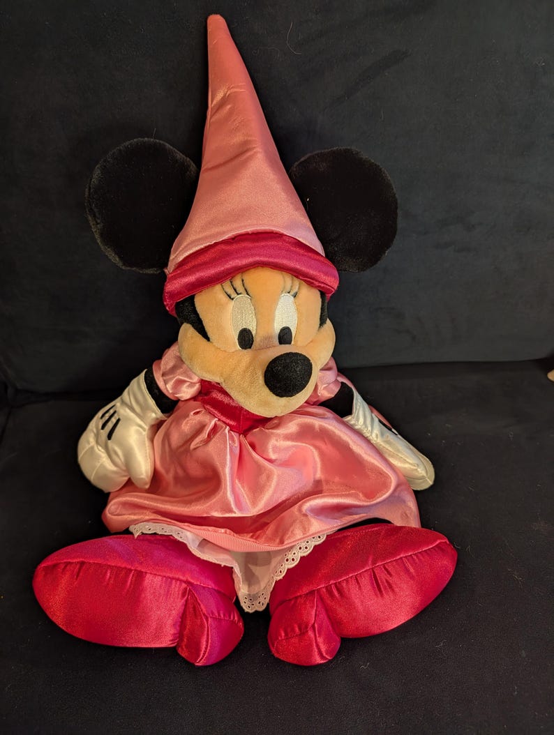 Peut inclure: Poup&eacute;e en peluche Minnie Mouse v&ecirc;tue d'une robe en satin rose, d'un chapeau assorti et de grandes chaussures rouges. La poup&eacute;e a des oreilles noires, un visage bronz&eacute; et des gants blancs. La robe a une bordure en dentelle blanche.