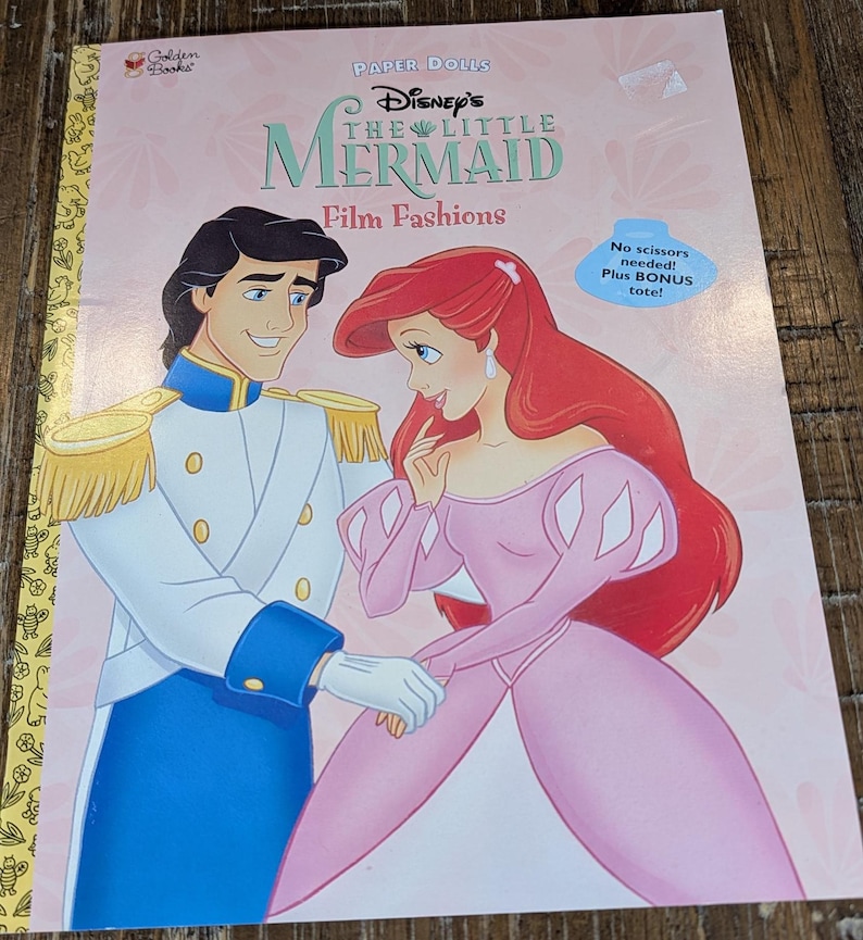May include: A children's book titled "Disney's The Little Mermaid Film Fashions" featuring Ariel and Prince Eric. The cover is pink with illustrations of the characters and text. Includes a bonus tote. The Golden Books logo is in the upper left corner.