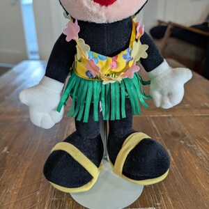 May include: A plush Minnie Mouse doll wearing a yellow floral top, green grass skirt, and yellow sandals. The doll has black fur, white gloves, and a red mouth. The doll is standing on a white stand.