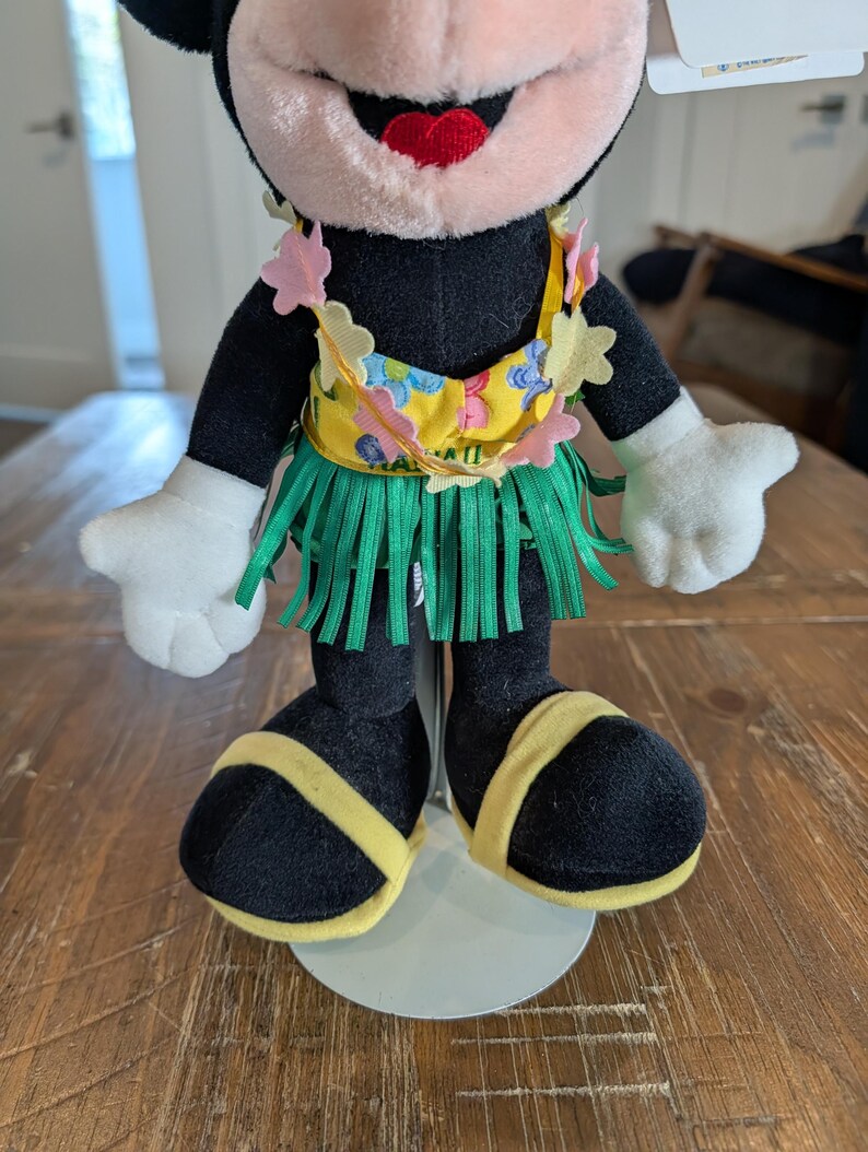 May include: A plush Minnie Mouse doll wearing a yellow floral top, green grass skirt, and yellow sandals. The doll has black fur, white gloves, and a red mouth. The doll is standing on a white stand.