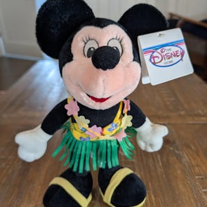 May include: A plush Minnie Mouse doll wearing a yellow and green hula skirt and a floral lei. The doll has black ears, a pink face, and a red mouth. A Disney Store tag is attached.