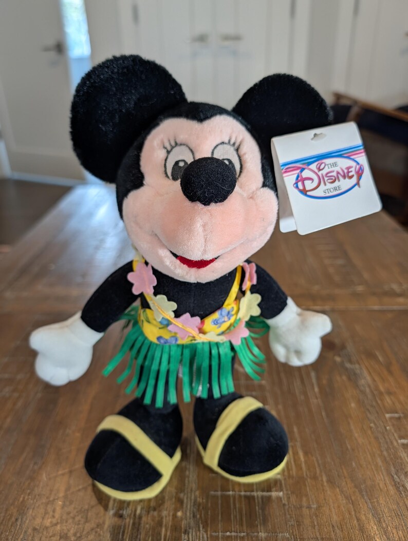May include: A plush Minnie Mouse doll wearing a yellow and green hula skirt and a floral lei. The doll has black ears, a pink face, and a red mouth. A Disney Store tag is attached.