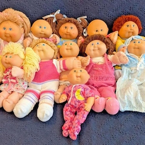 May include: A collection of ten Cabbage Patch Kids dolls with various hair colors, including blonde, brown, and auburn, wearing assorted outfits such as pink overalls, floral dresses, and patterned pajamas, all featuring signature soft bodies and vinyl heads.