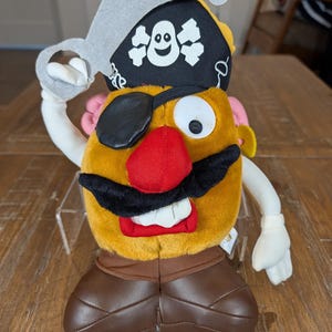 May include: A plush toy potato dressed as a pirate, featuring a black eye patch, a red nose, and a black mustache. It wears a pirate hat with a skull and crossbones and holds a gray sword. The toy has brown shoes and white gloves.