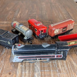 May include: A collection of vintage metal toy train cars including a black New York Central locomotive, a silver tank car, a red caboose, a red boxcar, and a green and red open-top freight car. The black locomotive features red and silver detailing.