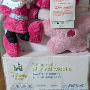 May include: A Disney Baby musical mobile featuring Minnie Mouse plush figures in pink and black outfits. The packaging includes the text "Sitting Pretty Musical Mobile" and safety warnings. The mobile is designed for babies from birth to 5 months.