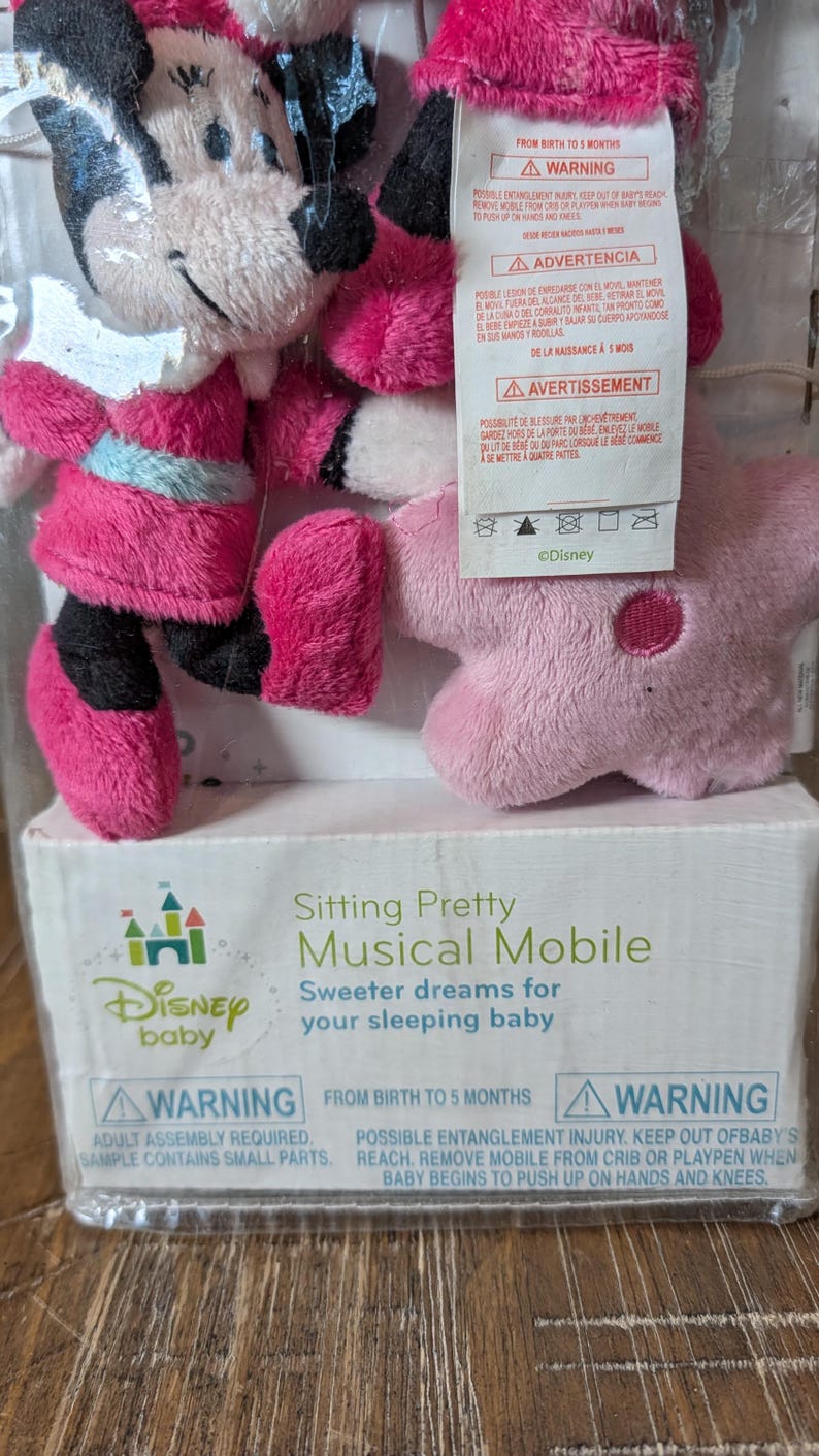 May include: A Disney Baby musical mobile featuring Minnie Mouse plush figures in pink and black outfits. The packaging includes the text "Sitting Pretty Musical Mobile" and safety warnings. The mobile is designed for babies from birth to 5 months.