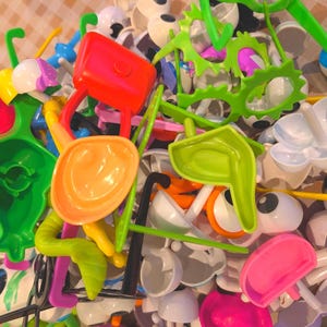 May include: A colorful assortment of plastic toy parts, including green, orange, pink, and yellow pieces. The image shows various shapes, such as eye-shaped components, handles, and other geometric forms, creating a playful and vibrant display.
