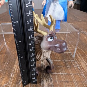 May include: A brown and tan plastic toy reindeer, approximately 5 inches tall, with large antlers and a friendly expression. A black ruler is placed next to the toy. Two dolls are in the background.