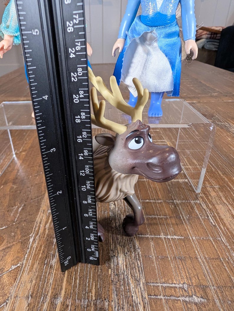 May include: A brown and tan plastic toy reindeer, approximately 5 inches tall, with large antlers and a friendly expression. A black ruler is placed next to the toy. Two dolls are in the background.