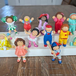 May include: A collection of vintage Cabbage Patch Kids figurines. The figurines are in various outfits and poses, including a cheerleader, a baby with a pacifier, and a baseball player. The color palette includes red, yellow, blue, and pink.
