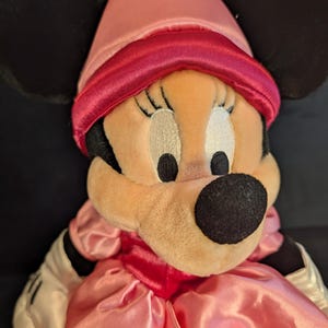 Peut inclure: Poup&eacute;e en peluche Minnie Mouse v&ecirc;tue d'une robe en satin rose et d'un chapeau pointu assorti avec une bande rouge. La poup&eacute;e a des oreilles noires, un nez noir et des gants blancs.