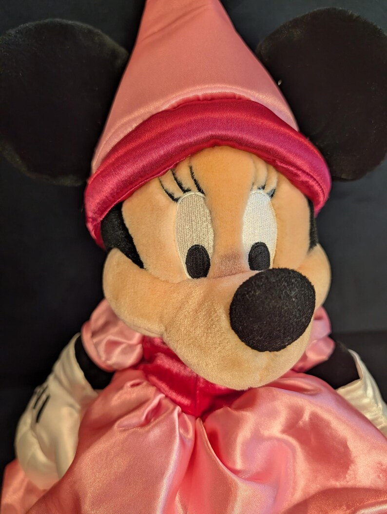 Peut inclure: Poup&eacute;e en peluche Minnie Mouse v&ecirc;tue d'une robe en satin rose et d'un chapeau pointu assorti avec une bande rouge. La poup&eacute;e a des oreilles noires, un nez noir et des gants blancs.