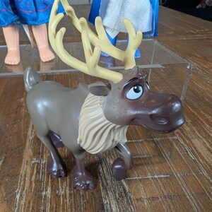 May include: A brown and tan plastic toy reindeer with large antlers and a white ruffled collar. The reindeer has a sad expression and is posed in a walking position. Other dolls are visible in the background.