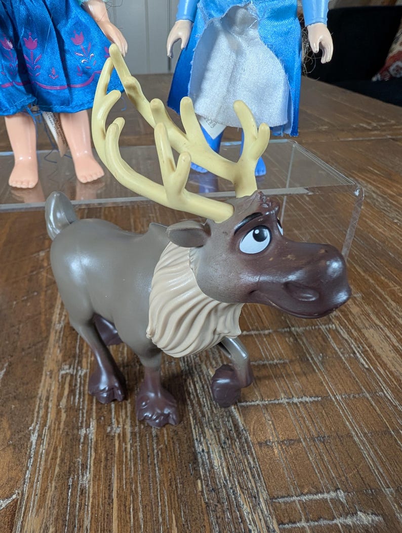 May include: A brown and tan plastic toy reindeer with large antlers and a white ruffled collar. The reindeer has a sad expression and is posed in a walking position. Other dolls are visible in the background.
