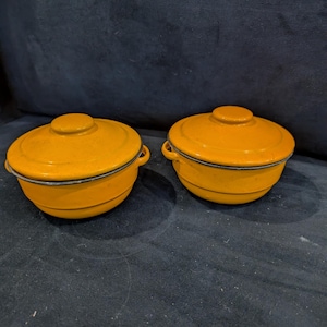 May include: Two yellow enamelware casserole dishes with lids. The dishes have black rims and are sitting on a dark gray surface.