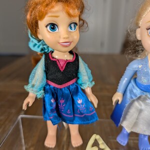 May include: A doll of a character with red hair, blue eyes, and a blue dress. The doll has a black top with pink trim and a blue skirt with a floral design. The doll is barefoot and has a smiling expression. Another doll is partially visible.