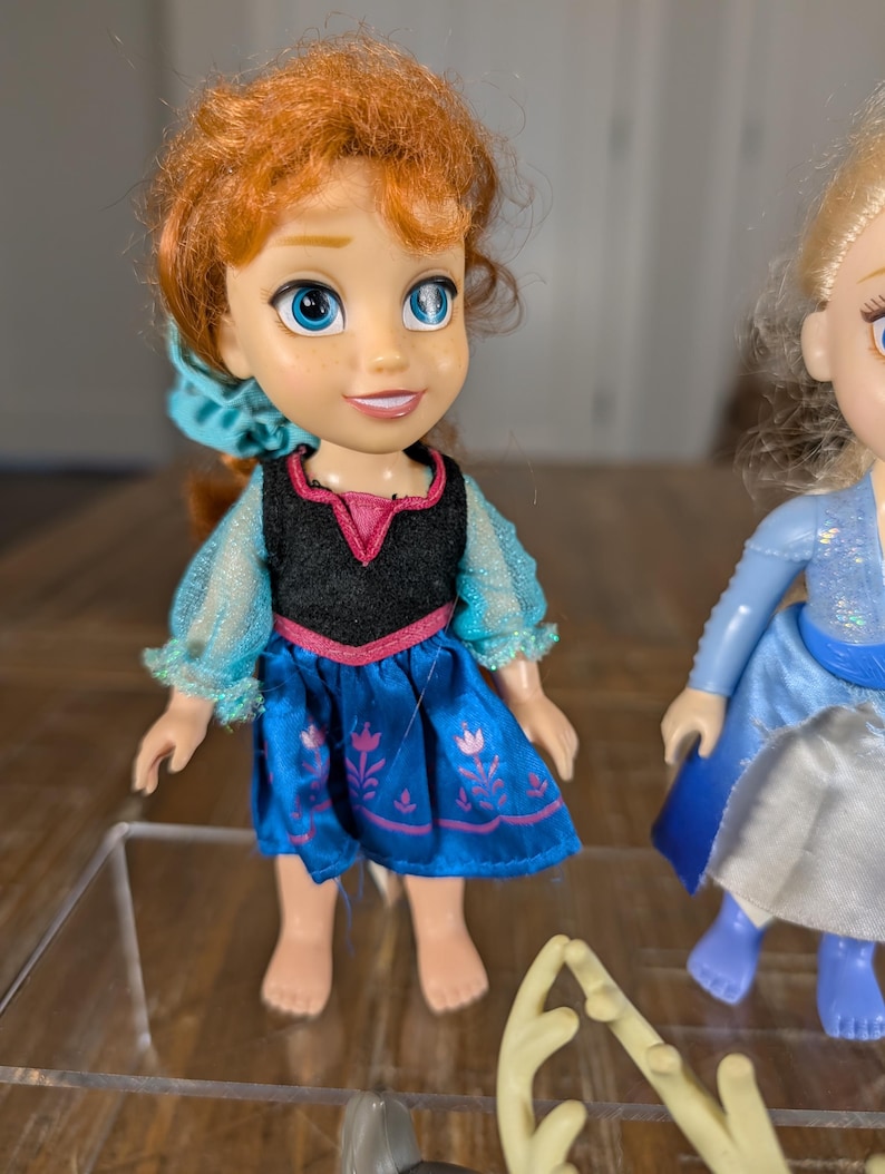 May include: A doll of a character with red hair, blue eyes, and a blue dress. The doll has a black top with pink trim and a blue skirt with a floral design. The doll is barefoot and has a smiling expression. Another doll is partially visible.