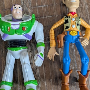 May include: Two plastic action figures representing a space ranger and a cowboy. The space ranger features a white, green, and purple suit with a purple hood. The cowboy wears a yellow plaid shirt, a cow-print vest, blue pants, and brown boots.