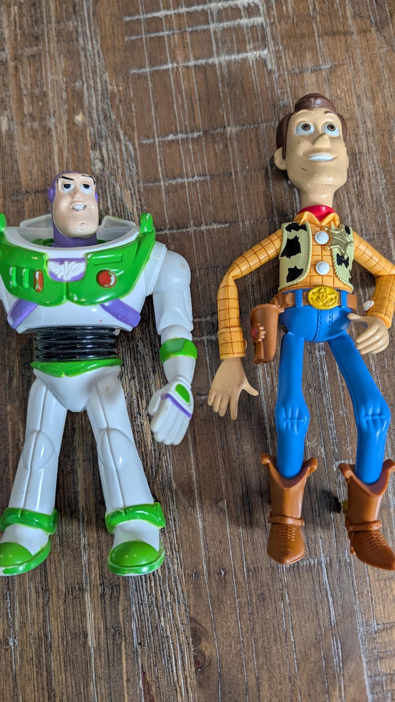 May include: Two plastic action figures representing a space ranger and a cowboy. The space ranger features a white, green, and purple suit with a purple hood. The cowboy wears a yellow plaid shirt, a cow-print vest, blue pants, and brown boots.