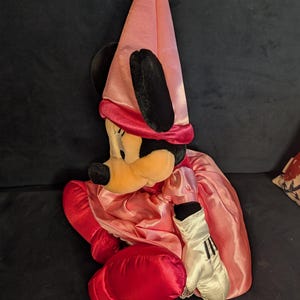 Peut inclure: Poup&eacute;e en peluche Minnie Mouse v&ecirc;tue d'une robe en satin rose et d'un chapeau pointu assorti. La poup&eacute;e a des oreilles noires, un visage jaune et des gants rouges. La poup&eacute;e est assise sur une surface bleu fonc&eacute;.