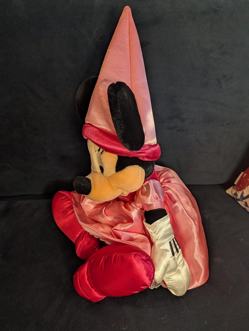 Peut inclure: Poup&eacute;e en peluche Minnie Mouse v&ecirc;tue d'une robe en satin rose et d'un chapeau pointu assorti. La poup&eacute;e a des oreilles noires, un visage jaune et des gants rouges. La poup&eacute;e est assise sur une surface bleu fonc&eacute;.