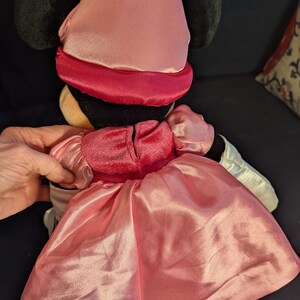 Peut inclure: Poup&eacute;e en peluche Minnie Mouse, vue de dos, v&ecirc;tue d'une robe en satin rose et d'un chapeau assorti. La poup&eacute;e a des oreilles noires et un bandeau rouge autour de la t&ecirc;te. La robe a une jupe ample et des manches bouffantes. La poup&eacute;e est sur une surface sombre.