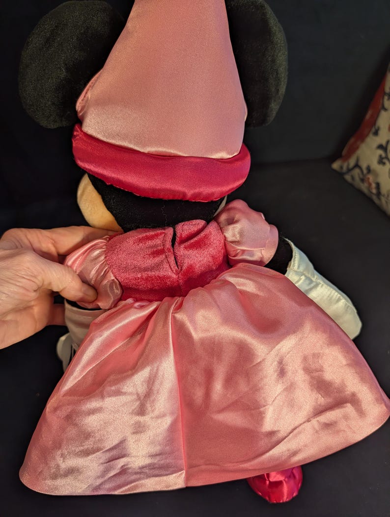 Peut inclure: Poup&eacute;e en peluche Minnie Mouse, vue de dos, v&ecirc;tue d'une robe en satin rose et d'un chapeau assorti. La poup&eacute;e a des oreilles noires et un bandeau rouge autour de la t&ecirc;te. La robe a une jupe ample et des manches bouffantes. La poup&eacute;e est sur une surface sombre.