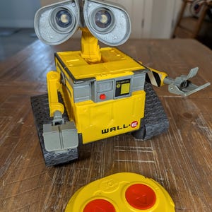 May include: A yellow and gray WALL-E toy robot with large, binocular-like eyes and a remote control. The robot has a box-shaped body, treads, and articulated arms. The remote is yellow with red buttons. The robot is on a wooden surface.