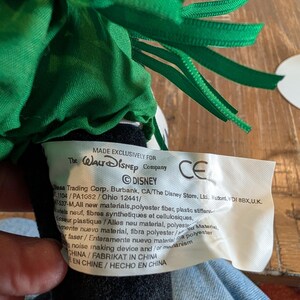 May include: A close-up of a plush toy with a green ribbon and a tag that reads "MADE EXCLUSIVELY FOR The Walt Disney Company". The tag also includes the Disney logo and other text.