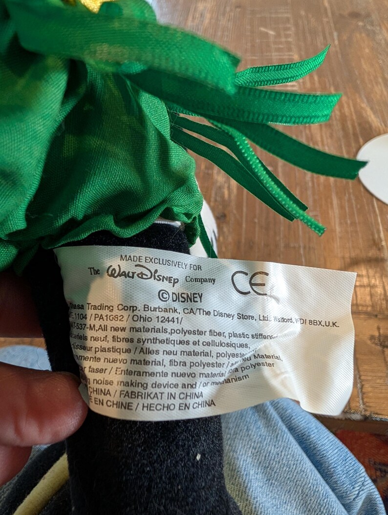May include: A close-up of a plush toy with a green ribbon and a tag that reads "MADE EXCLUSIVELY FOR The Walt Disney Company". The tag also includes the Disney logo and other text.
