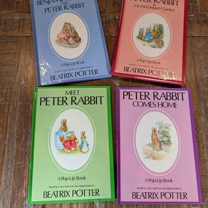 May include: Four Beatrix Potter pop-up books with colorful covers: light blue Benjamin Bunny Visits Peter Rabbit, pink Peter Rabbit in Mr. McGregor's Garden, green Meet Peter Rabbit, and purple Peter Rabbit Comes Home, all featuring illustrations.